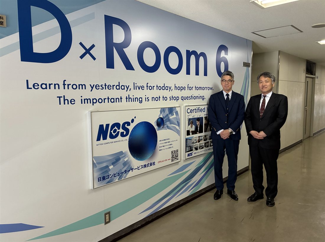 計算機実習室の壁面に大きく掲げられた「D×Room 6」のロゴと、その前に立つ大学・日東コンピューターサービス株式会社の担当者