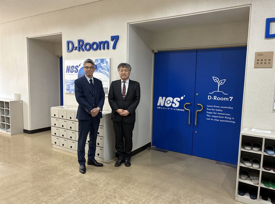 情報処理実習室Ⅲの入口扉に掲げられた「D×Room 7」のロゴと、その前に立つ大学・日東コンピューターサービス株式会社の担当者