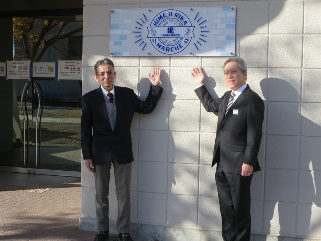 大学会館の外壁に掲げられた「HIMEJI RIKA MARCHE」のロゴプレートと、その前に立つ大学・ヒメジ理化株式会社の担当者