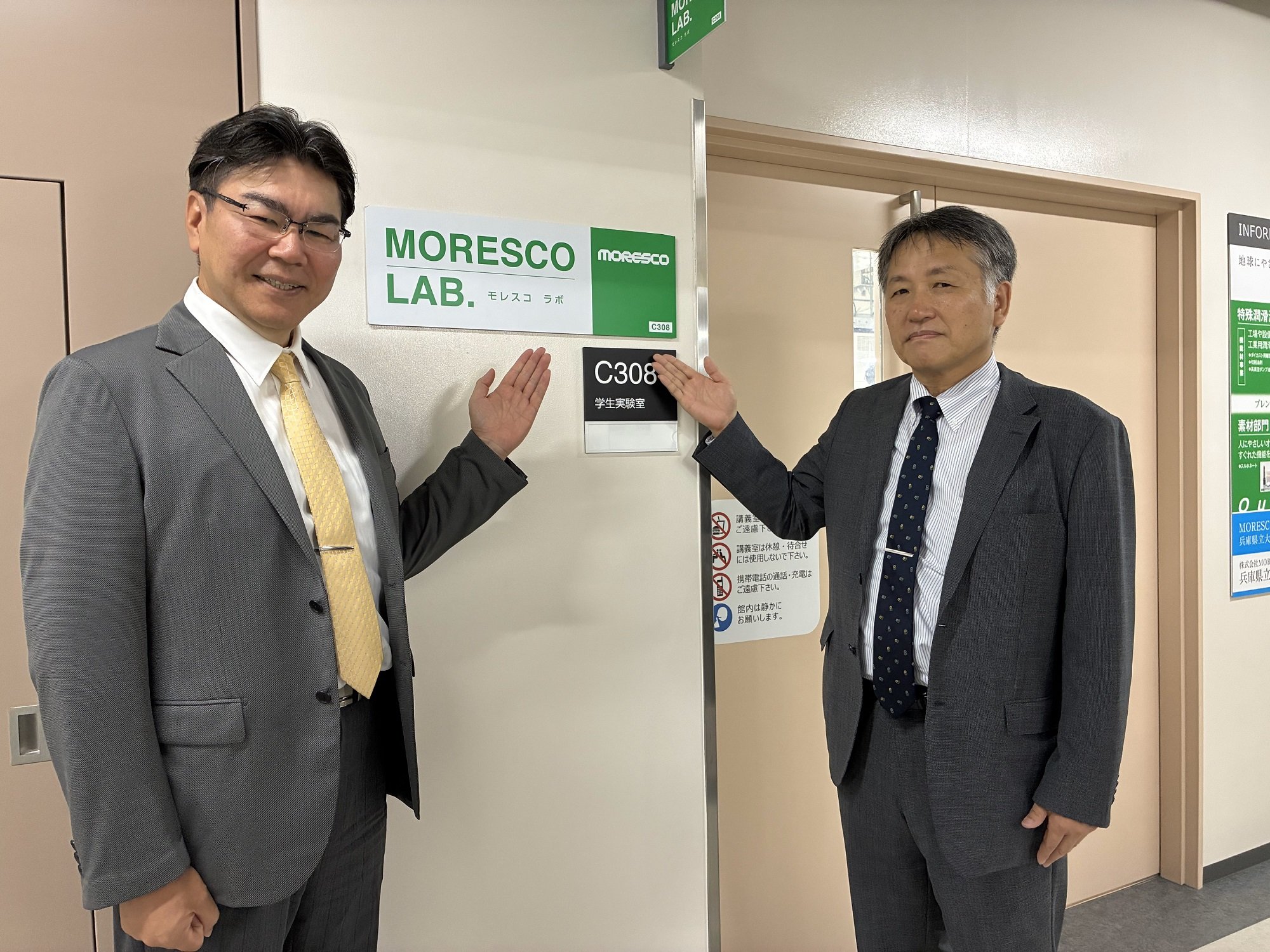 C308学生実験室の入口に掲げられた「MORESCO LAB.」の表示板と、その前に立つ大学・株式会社MORESCOの関係者