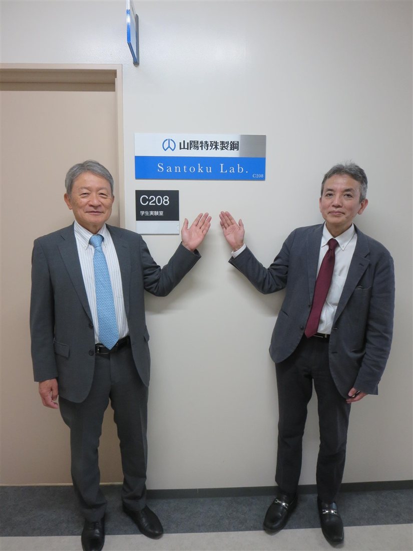 C208学生実験室の入口に掲げられた「山陽特殊製鋼 Santoku Lab. C208」の表示板と、その前に立つ大学・山陽特殊製鋼株式会社の担当者