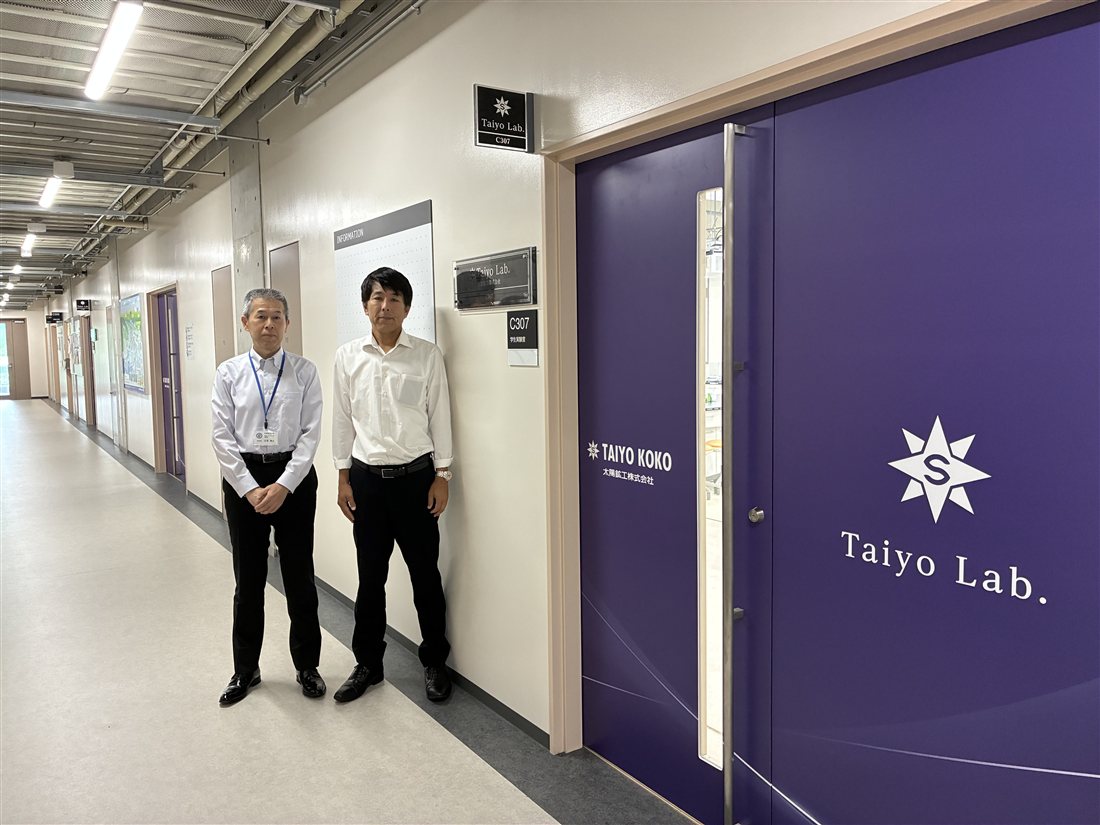 紫色の扉に「Taiyo Lab.」のロゴが掲げられたC307学生実験室の入口と、その前に立つ大学・太陽鉱工株式会社の担当者